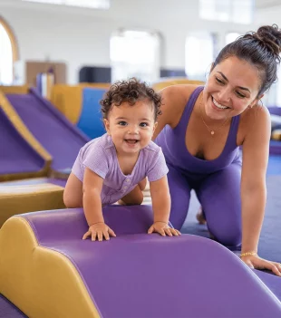 Toddler climbing a soft beam with a smiling coach beside her