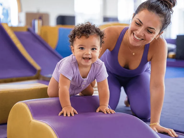 Toddler climbing a soft beam with a smiling coach beside her